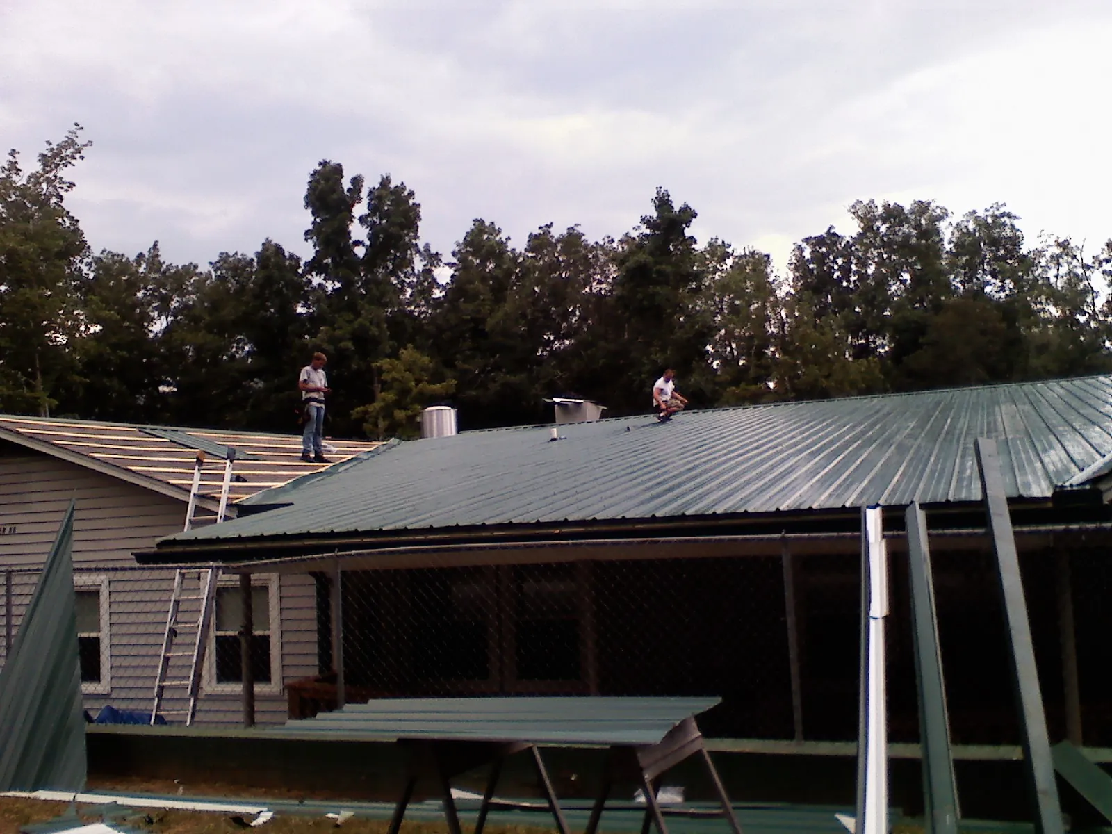 Two roofers installing green metal panels above a covered porch in University Park